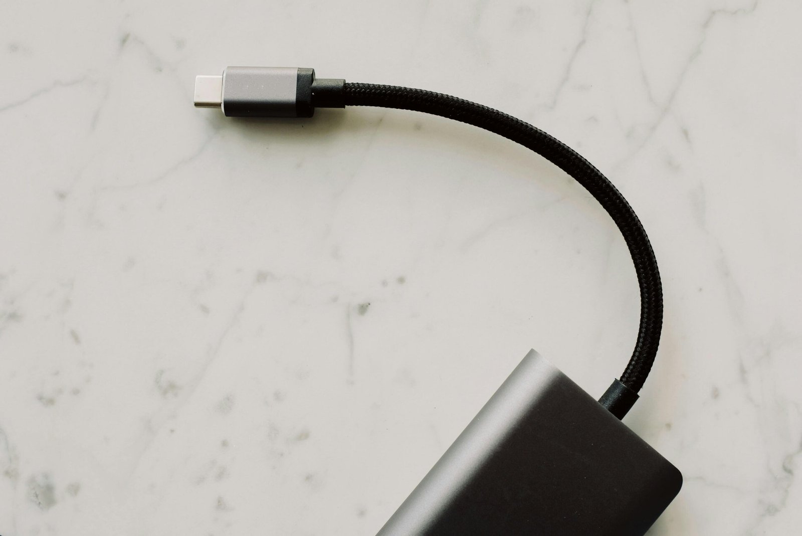 A close-up photo of a black USB-C cable with a braided sheath connected to a sleek, dark-colored external device, resting on a white marble surface.